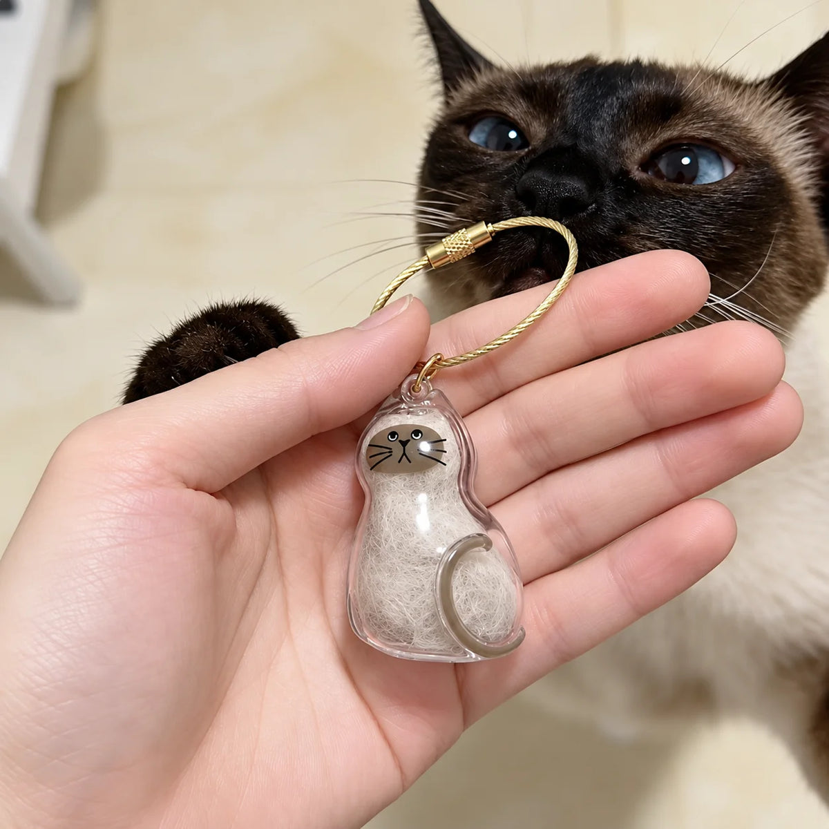 Main tenant un porte-clés PurrKey en forme de chat avec poils blancs, près d’un chat siamois